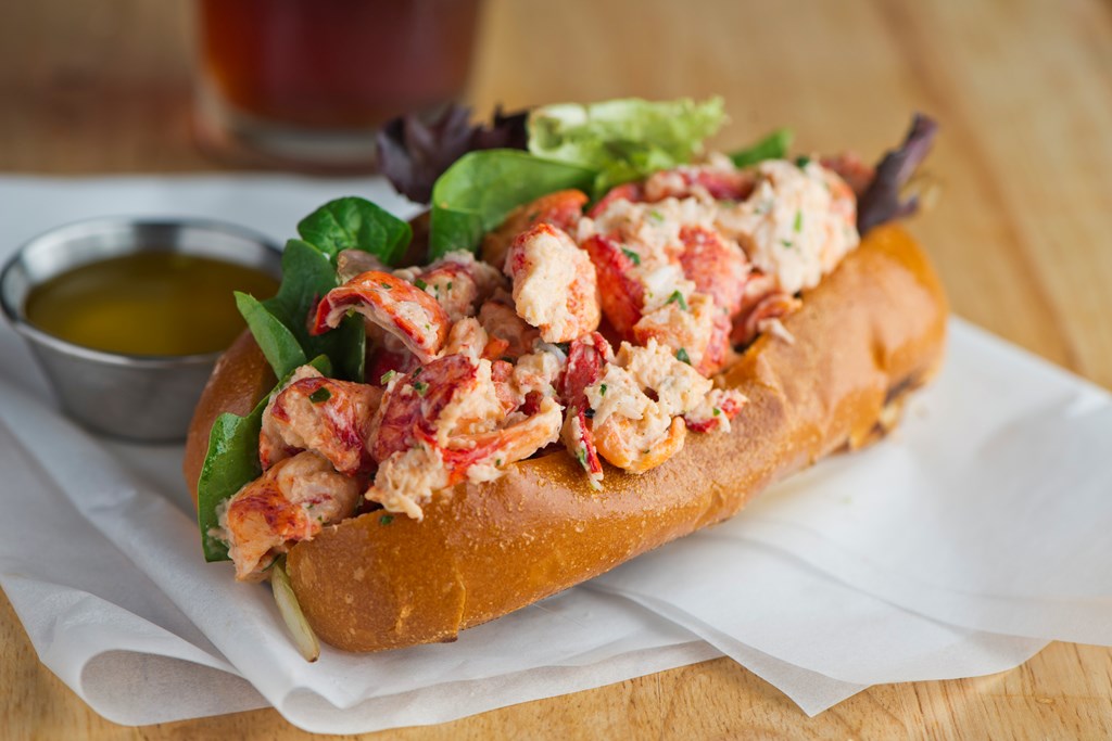 Lobster roll. Maine lobster mixed with mayo, celery, onions, garlic, scallions, chives, lemon juice. Lobster roll on toasted hotdog bun w/ lettuce, tomato, garlic mayo seasoned salt & pepper. Classic American restaurant or diner lunch sandwich favorite.