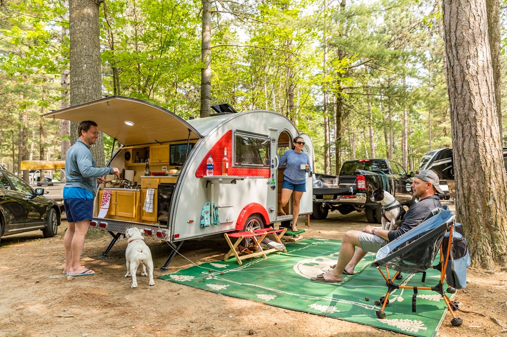 Three campers gather around a small travel trailer at a KOA campground.