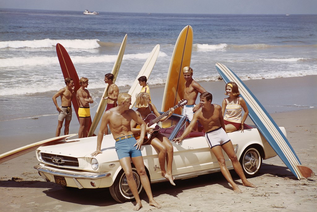 A vintage photo of a group of surfers on a beach with a convertible Ford Mustang.