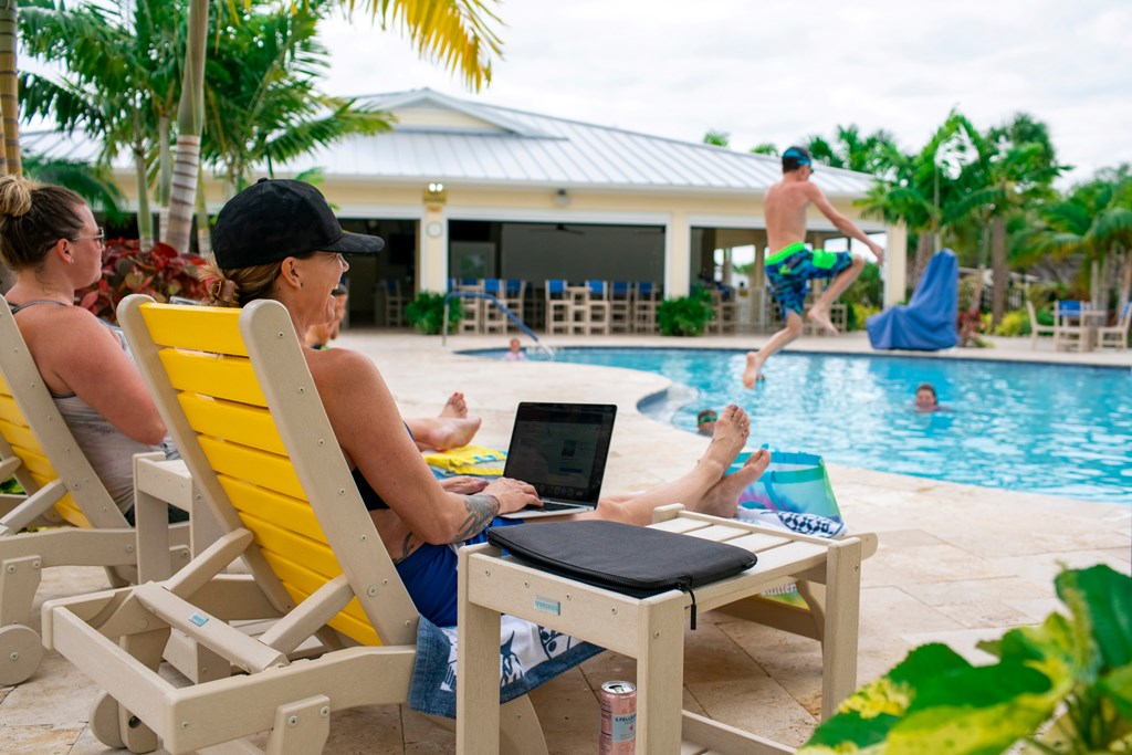 A woman on her laptop lounges by the pool as kids jump in.